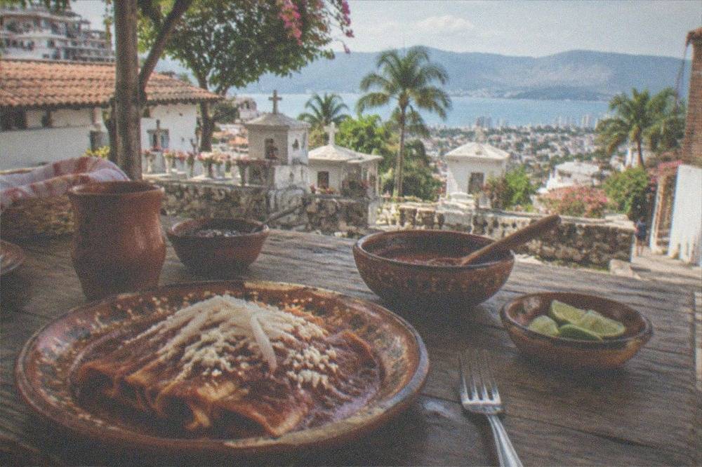 A rustic wooden table set with enchiladas, limes, and clay dishes ready for lunch