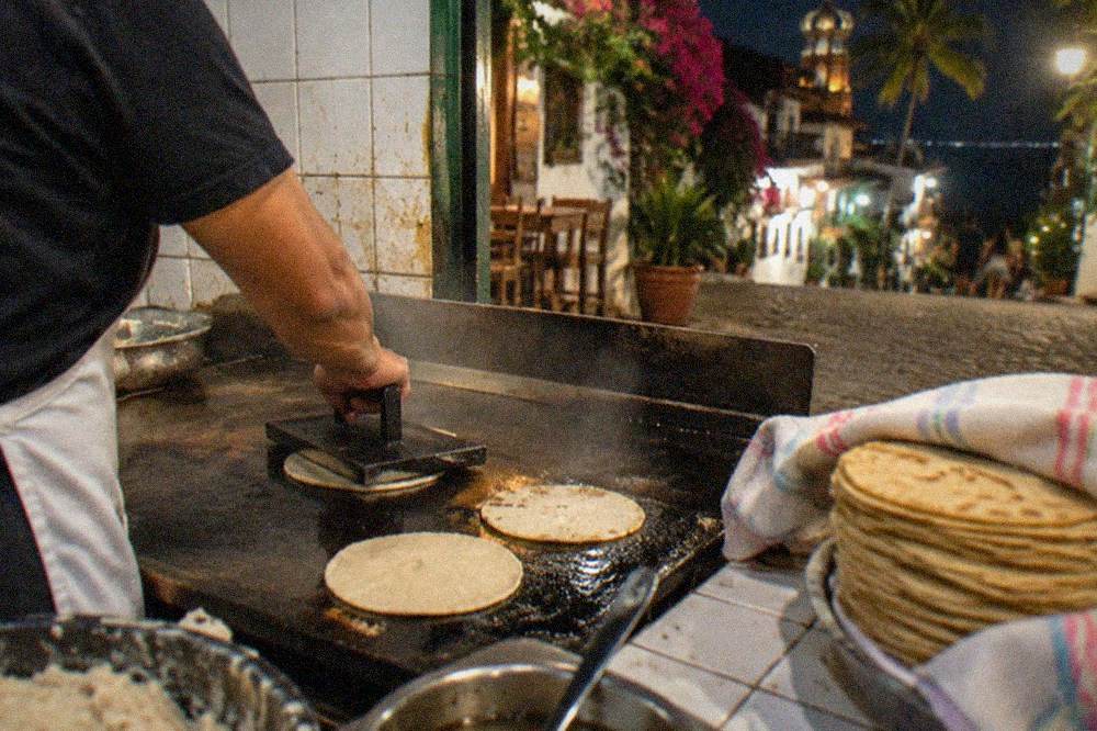 A cook pressing fresh masa tortillas on a hot comal with steam rising