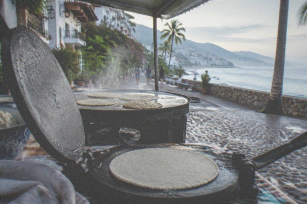 A vendor's hand preparing a corn tortilla on a hot griddle at a street stand