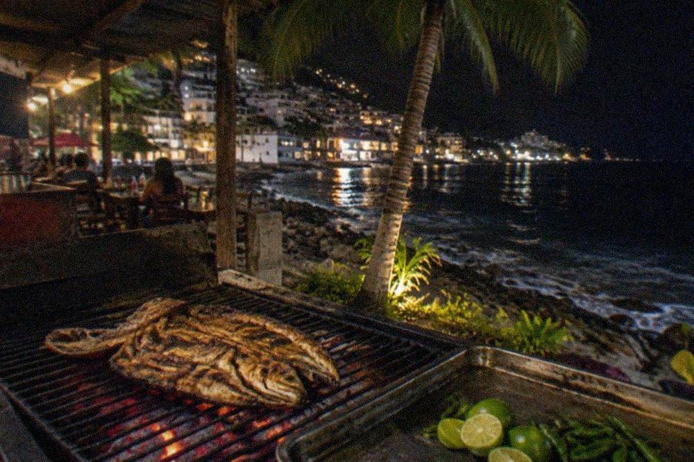 Whole grilled fish split open on a charcoal grill beside limes and chiles