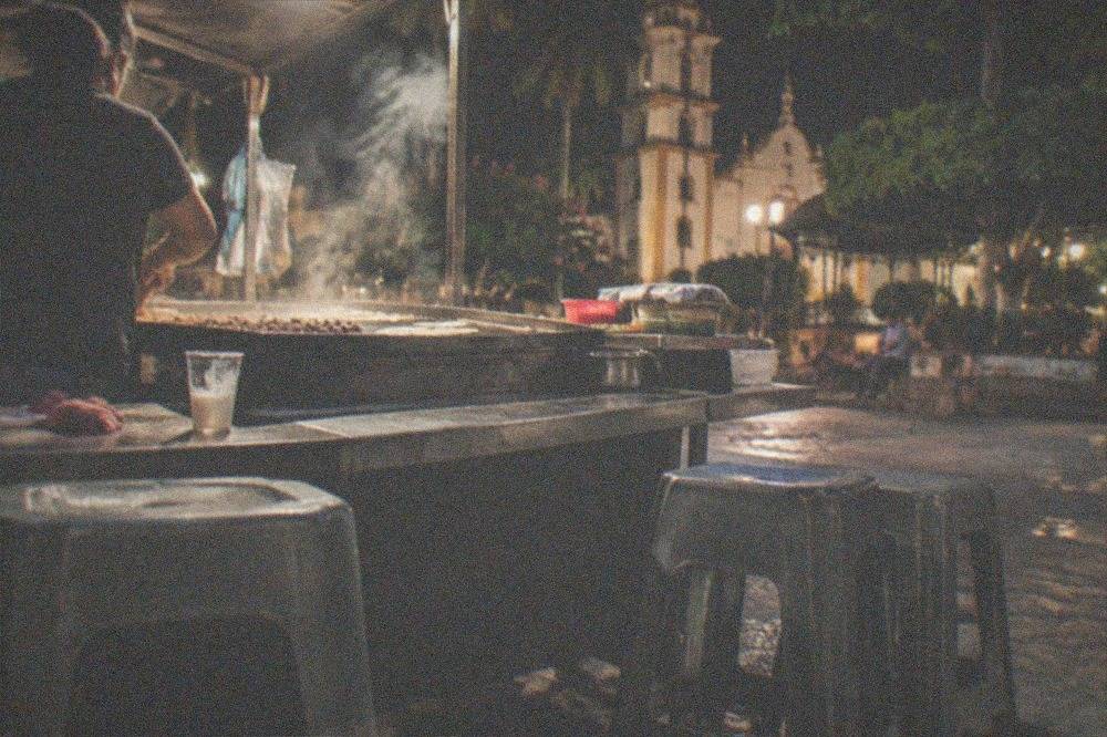 A lit taco stand at night with steam rising from a griddle and stools along the counter
