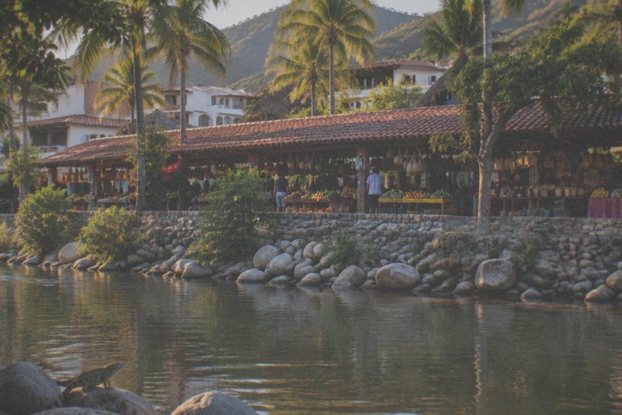 Covered market stalls with fresh fruit and hanging baskets beside a river in Puerto Vallarta