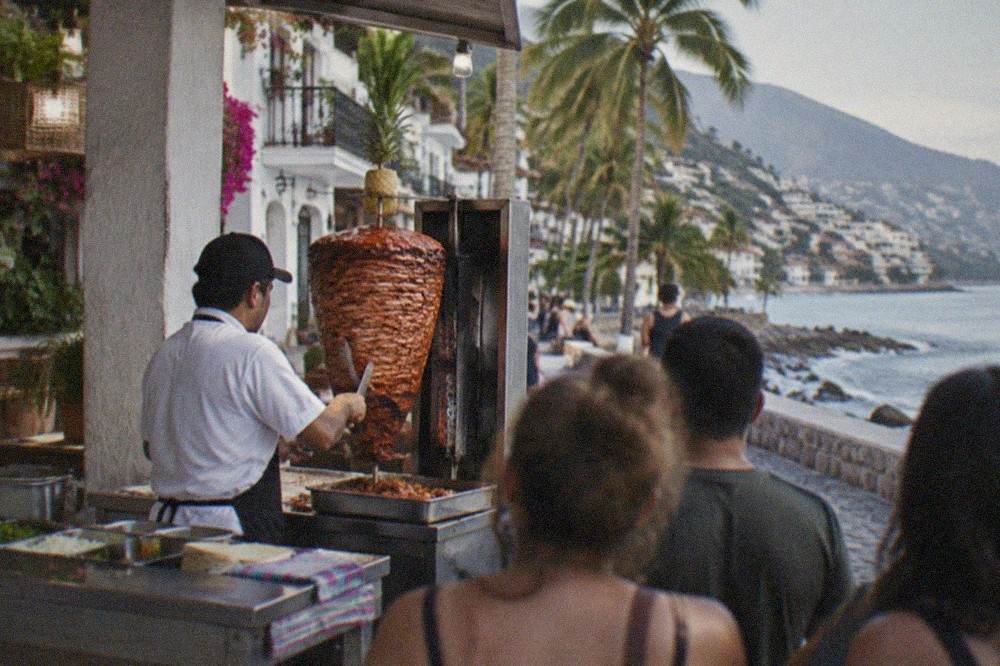 Al pastor tacos being carved from a vertical spit at a busy open-air taqueria counter