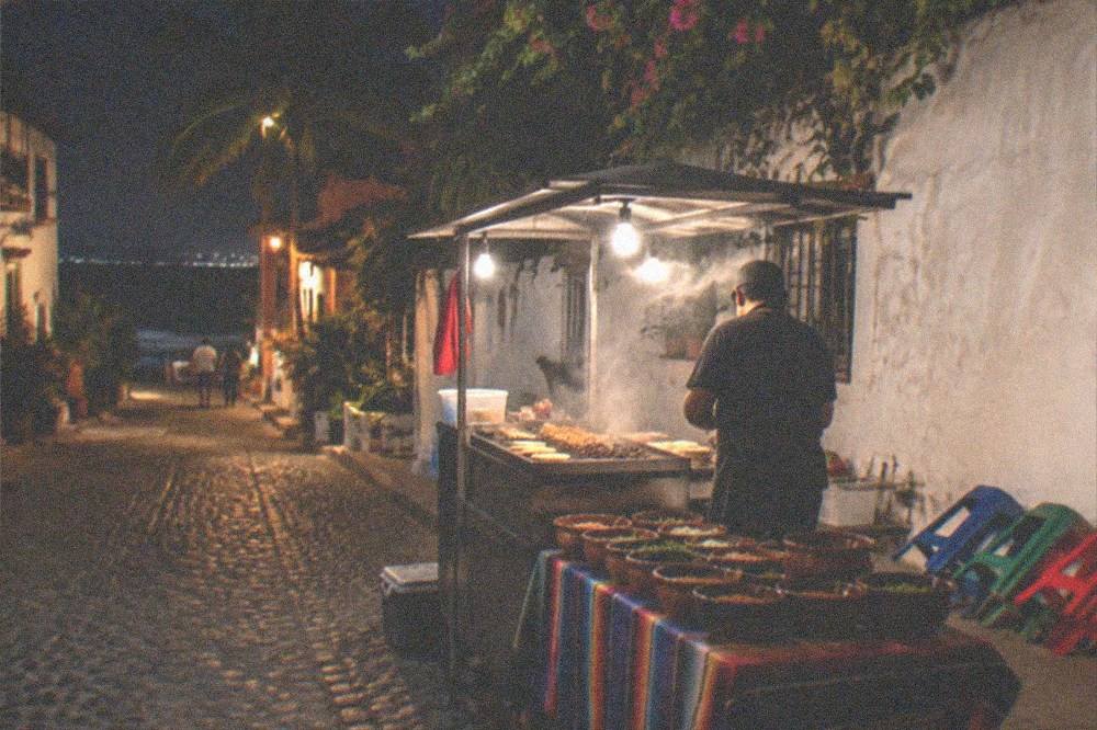 Street taco stand at night with griddle, salsas in small bowls and a row of stools