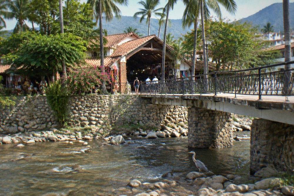Pedestrian footbridge over a shallow river leading to a market building among palm trees