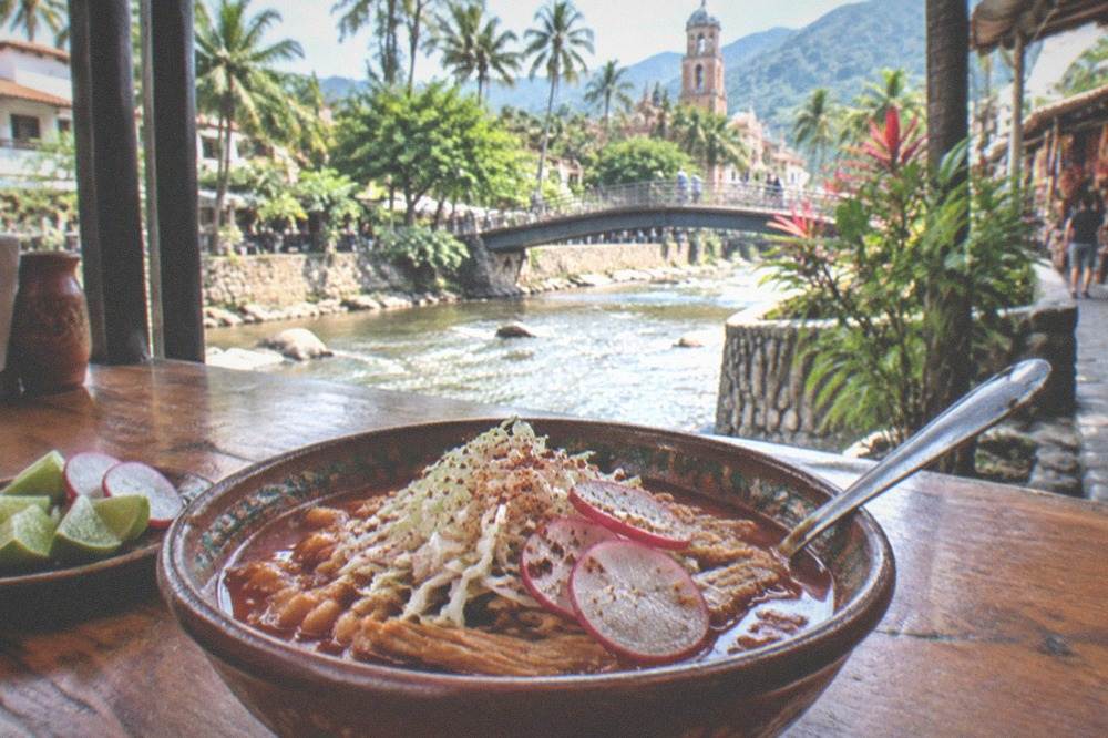 Bowl of red pozole with cabbage, radish and lime on a worn wooden counter
