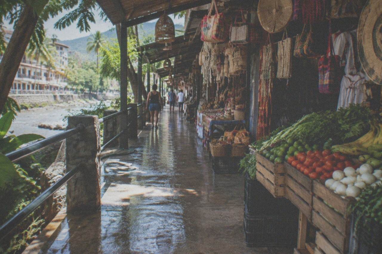 Market stalls with fresh produce and hanging crafts under a covered walkway beside a river