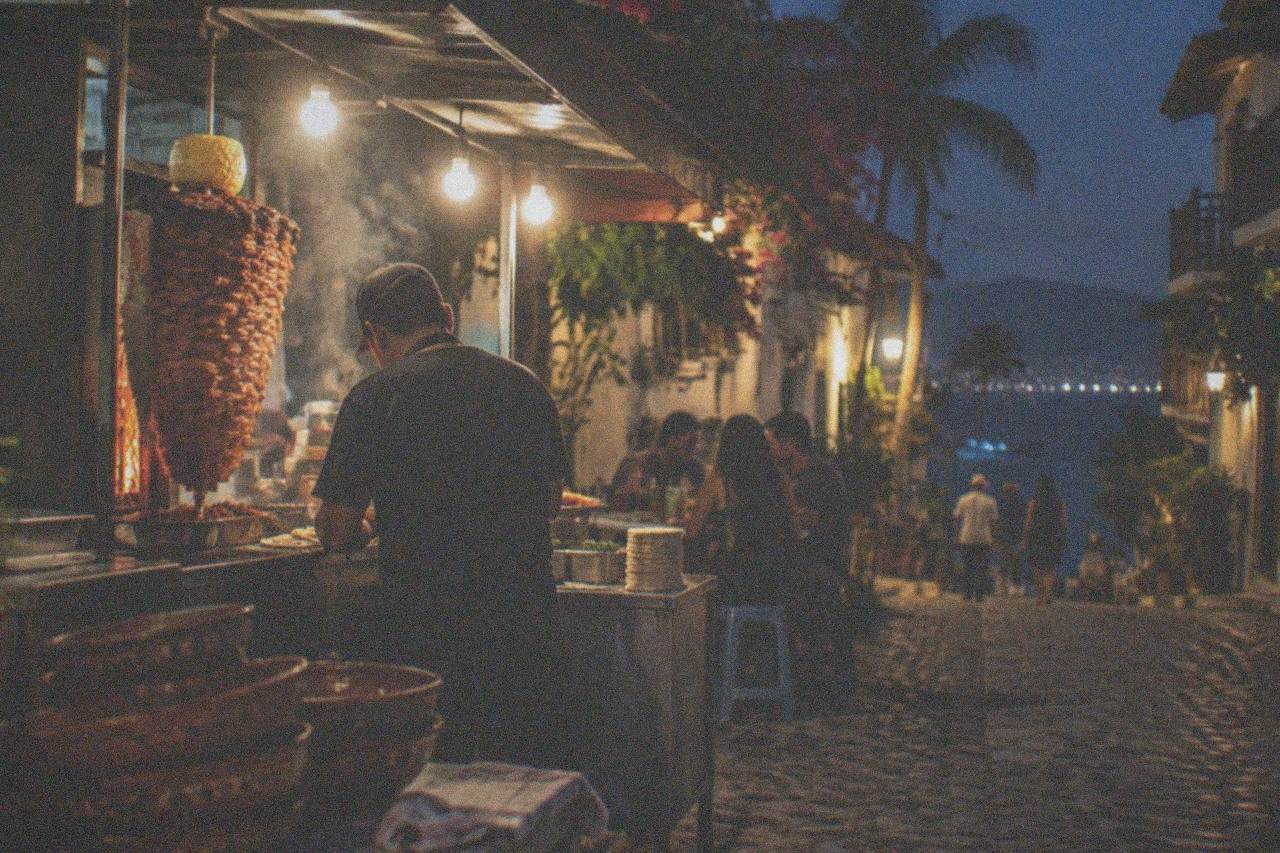 A busy evening taco stand with a grill full of al pastor meat in a Puerto Vallarta neighborhood