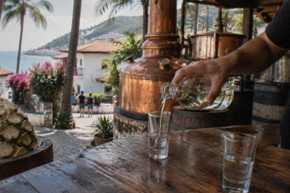 A guide pouring a small tasting of amber agave spirit into a caballito glass on a wooden table