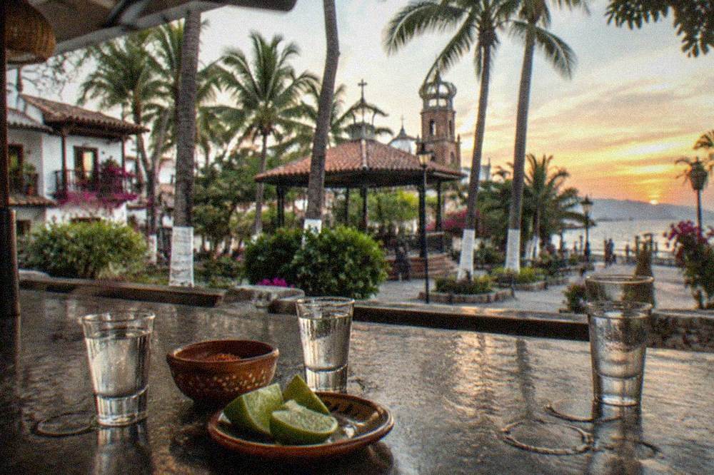 Three small tasting glasses of clear agave spirits beside sliced limes on a dark bar top