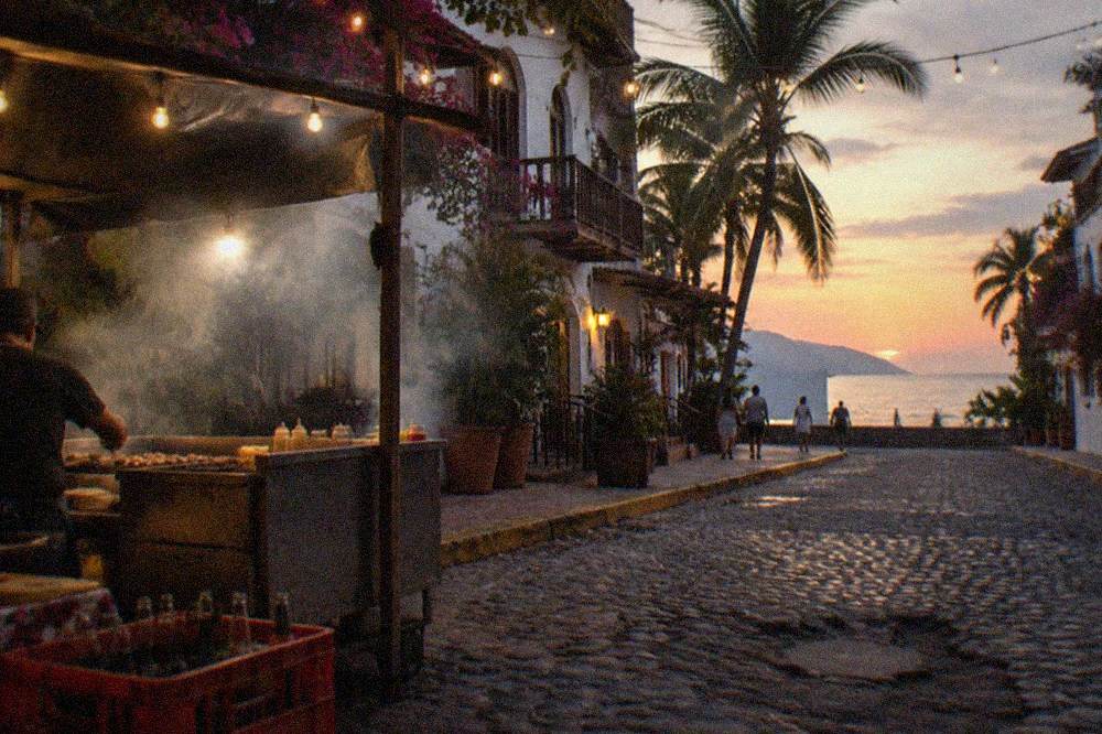 Open-air taco stand with grill smoke rising over a street corner at dusk