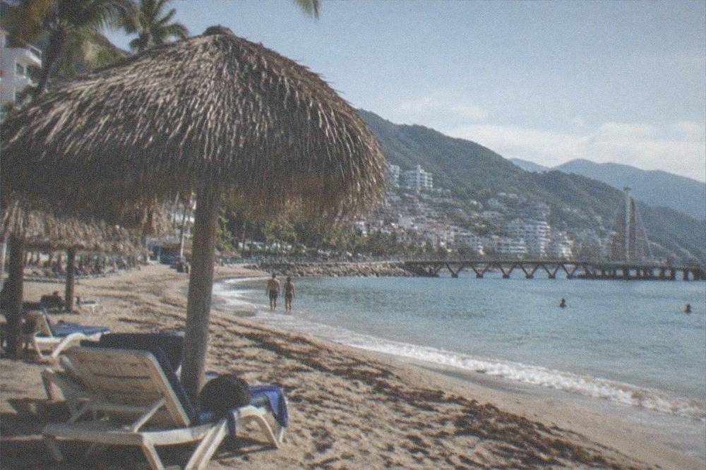 Long sandy beach with palm thatch umbrellas and a wooden pier extending into calm bay water