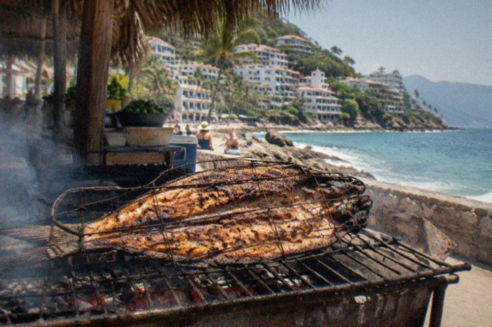 Charcoal grill with a split whole fish cooking over glowing coals at an outdoor seaside stall