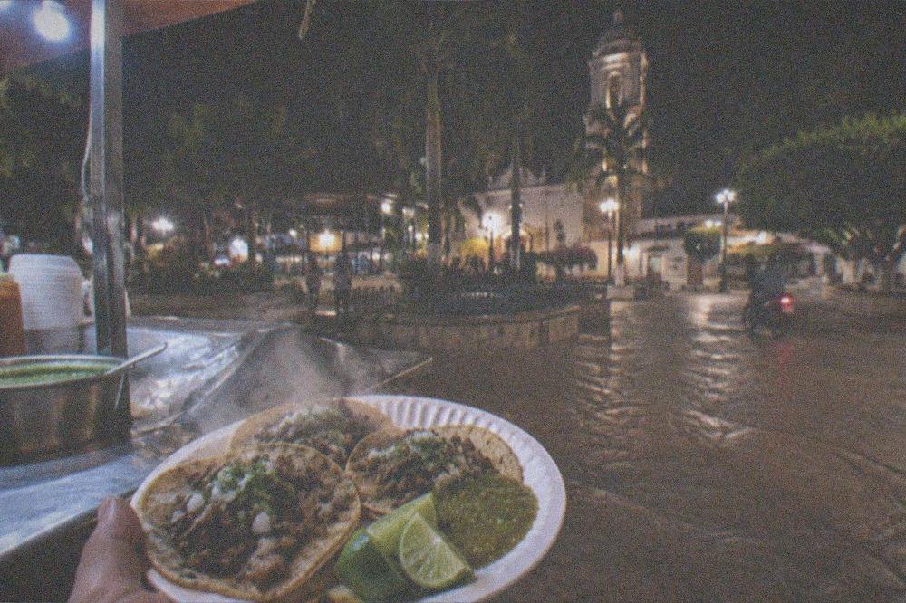 Hands holding a paper plate of street tacos with lime and green salsa at a night market stand